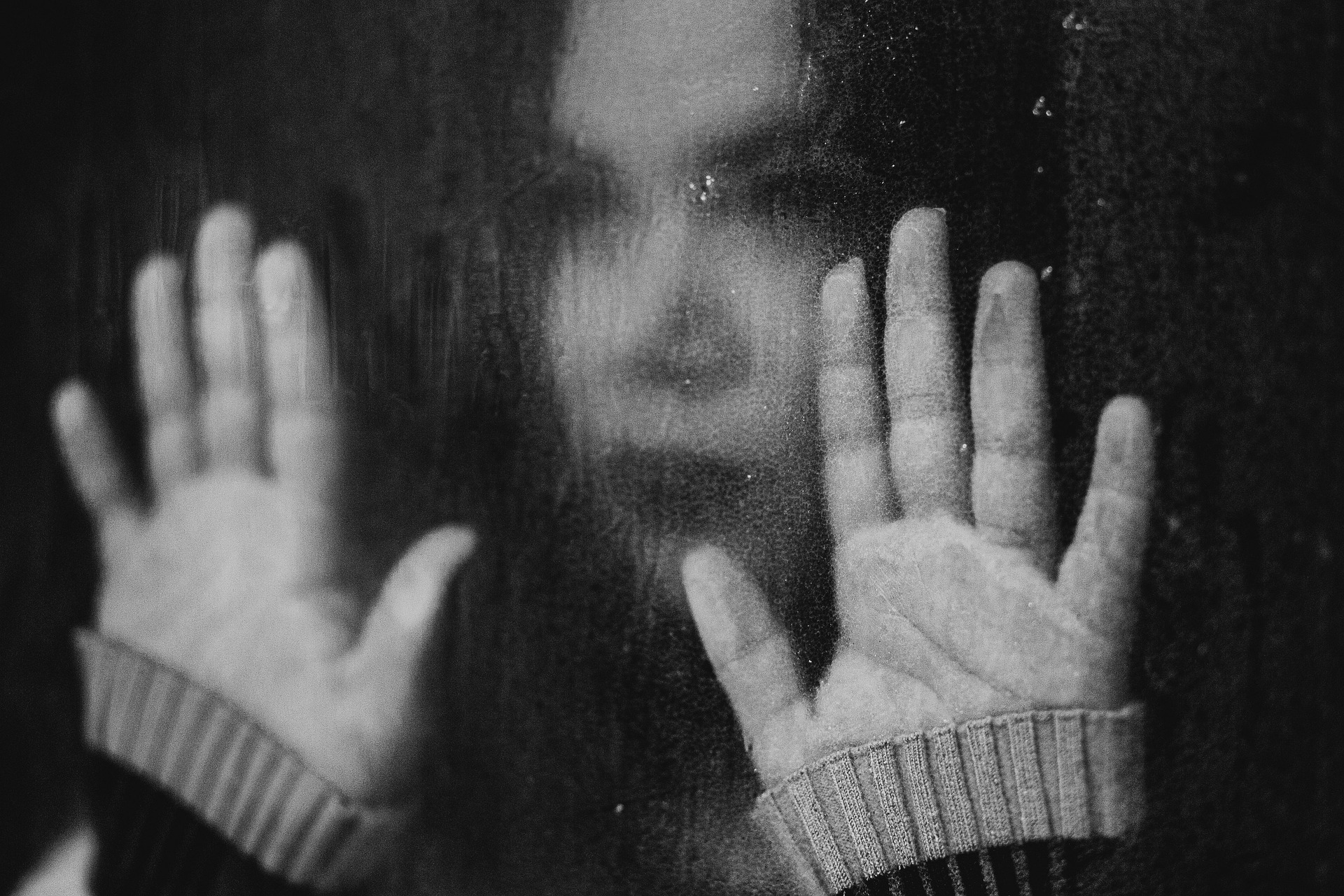 Young woman pressing her hands and face against a rain‑streaked window, looking out with a searching gaze.